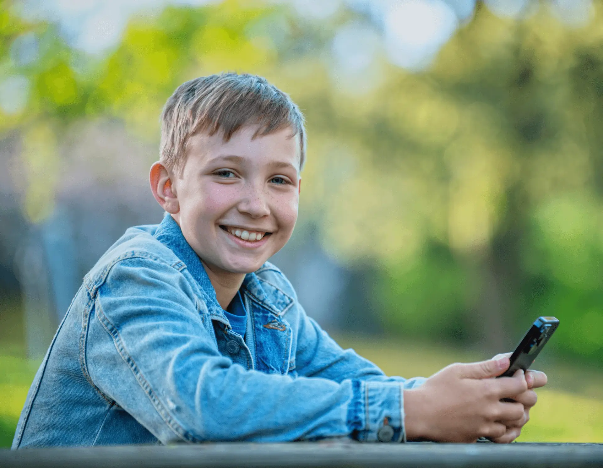 Smiling teen boy in denim jacket holding a phone outdoors
