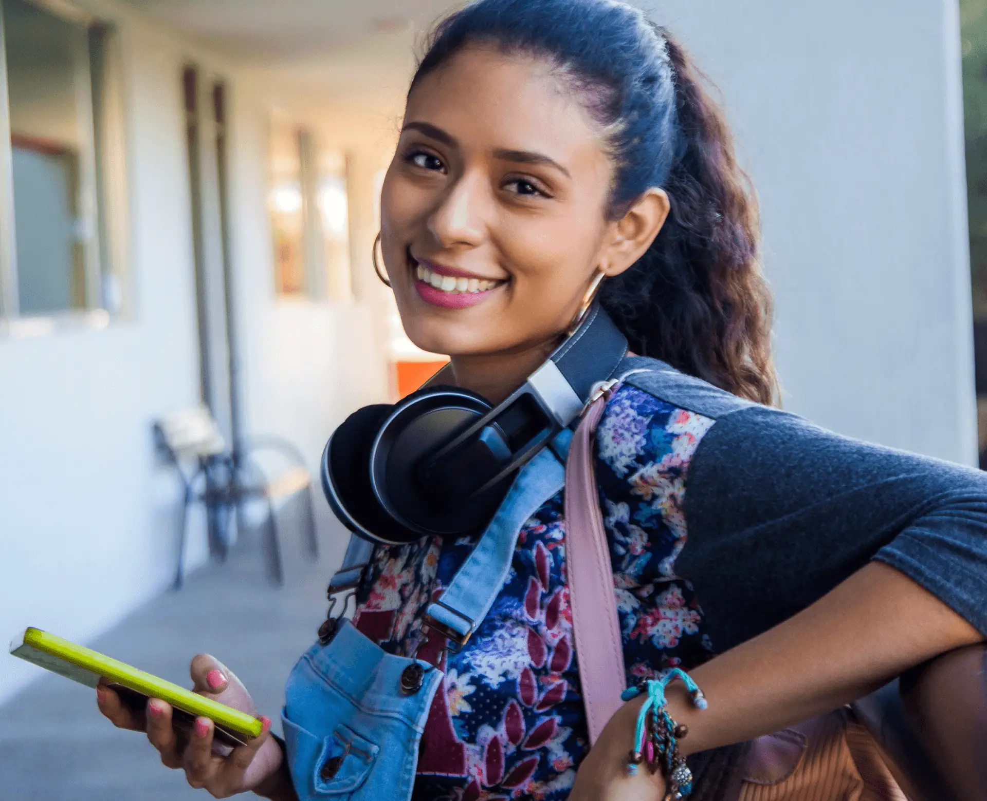 Smiling teen girl with ponytail, headphones around neck, holding phone and backpack strap