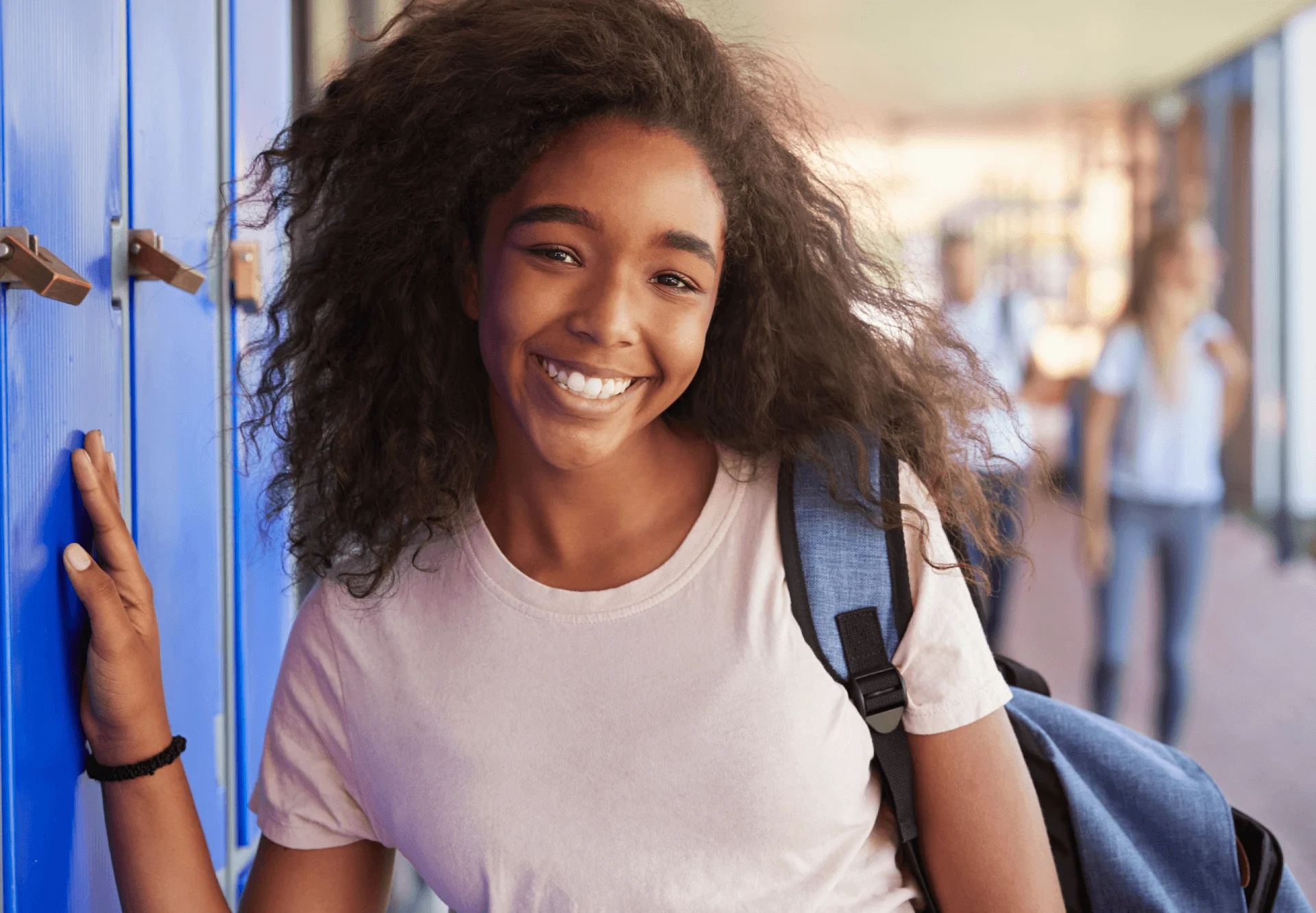 Smiling teen girl with backpack leaning against blue lockers in a school hallway