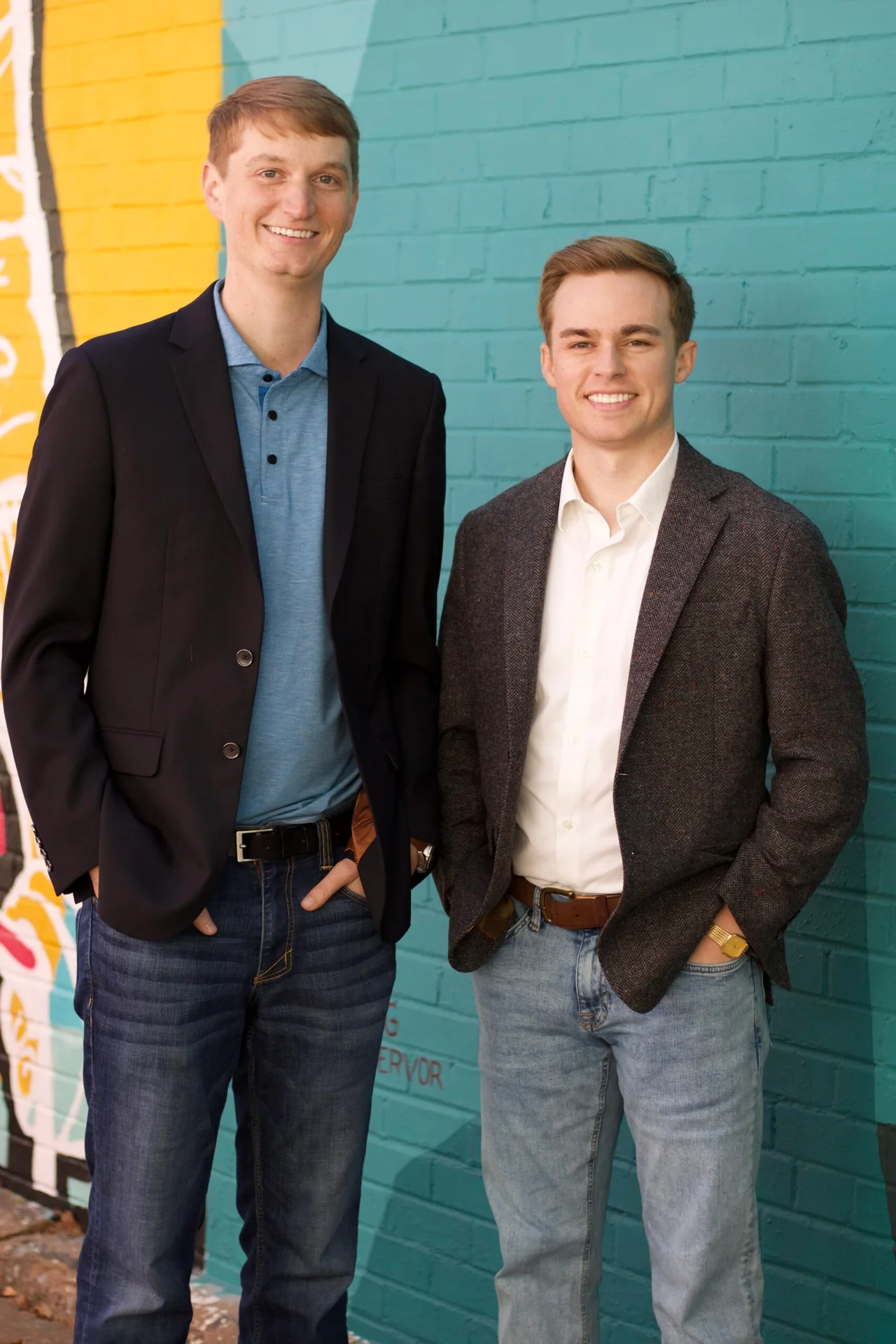 Two smiling young men in blazers and jeans posing in front of a teal brick wall mural